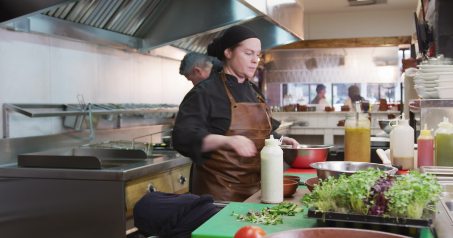 Side view of a Caucasian female cook working in a busy restaurant kitchen, preparing garnish in a bowl, her colleagues working in the background Royalty-Free Stock Footage #1045617868