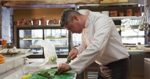 Side view of a Caucasian male chef working in a busy restaurant kitchen, slicing spring onions, female cook walking in the background