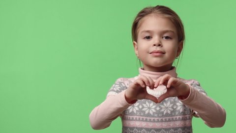 Video portrait of happy baby girl 6 years old in studio shows heart gesture by two hands, looking at camera. Sweet caucasian baby dressed in pink sweater stays on green chroma key background.