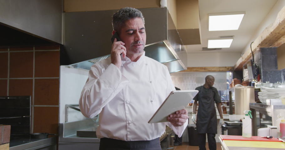 Front view of a Caucasian male chef working in a busy restaurant kitchen, holding a tablet computer and talking on the smartphone, male cook working in the background Royalty-Free Stock Footage #1045620658