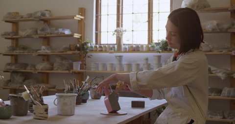 Side view of a young Caucasian female potter with auburn hair in a bob hairstyle sitting at a work table holding a pot and painting it with a brush in a pottery studio, with shelves and a window in