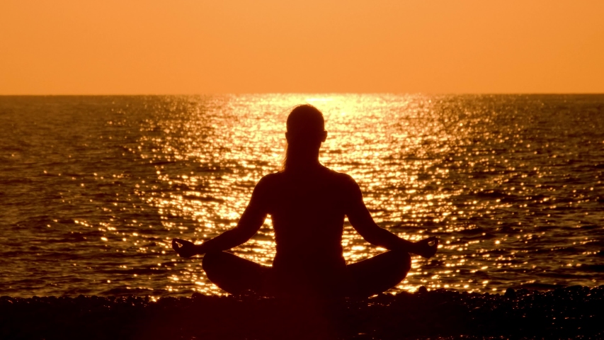Silhouette of woman is charged with energy of sun on seashore at yellow sunset. Girl meditates in lotus position on beach on background of sea at sunset. Royalty-Free Stock Footage #1045632916