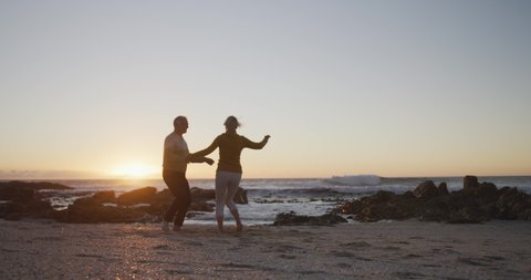 Front view of a senior Caucasian couple enjoying time in nature together, dancing on a beach at sunset, smiling, slow motion