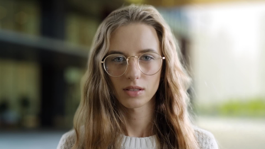 Portrait of serious clever girl in eyewear looking at a camera in a street background. A smart woman is smiling. Student with glasses outdoor