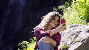 Young girl traveller take photo near mountain waterfall. Close-up portrait cheerful curly woman taking selfie, blogger posing and smiling on phone while recording vlog or showing video online