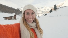 Happy young woman taking selfie portrait in the Swiss Alps in winter, mountains covered by snow, sunny day. girl video chatting on mobile phone sharing with friends family social media. travel winter 