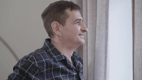 Side view close-up of adult Caucasian man looking out the window and smiling. Middle aged man enjoying morning at home. Guy leaving the shot. Leisure, lifestyle, joy. - Powered by Shutterstock - Get 15% off with code: PIKWIZARD15