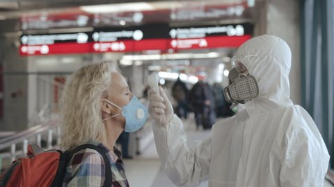 Controlling people's temperature and health at the entrance to airport, railway station or hypermarket. Medical worker in a protective suit screening passenger to check the Covid-19 symptoms