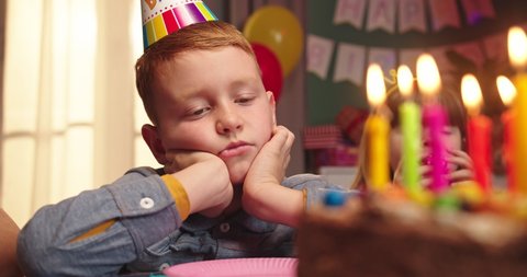 Close up of the teen little Caucasian bored boy in a conus sitting at the table in front of the birthday cake and blowing candles with unhappy face. Portrait.