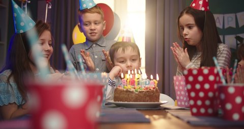 Happy little Caucasian boy celebrating a birthday - blowing candles and smiling at the camera while his friends blowing in horns and applausing.