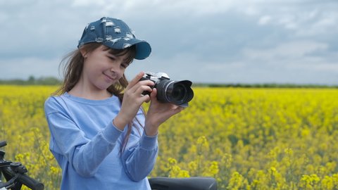 The child takes pictures. A nice little girl in a cap takes photos of yellow rape field.