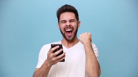 Handsome unshaven bearded young guy 20s wearing white t-shirt isolated over pastel blue background in studio. People sincere emotions lifestyle concept. Looking surprised wow, using mobile cell phone