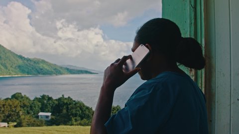 Melsisi, Pentecost Island / Vanuatu - May 10 2019: black female woman making a mobile cell phone call outside of the house in front of a beautiful sea shore landscape seascape
