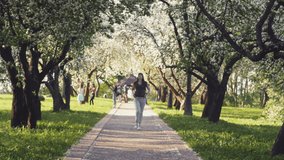 Young happy woman tourist is walking alone in spring sunny city park with blooming apple trees on background. Concept of beautiful girl weekend vacations. 4K slow motion video.