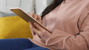 Young woman using rose gold tablet and seat on couch or sofa in jeans and pastel pink sweater. Woman using tablet for social network or shopping online.