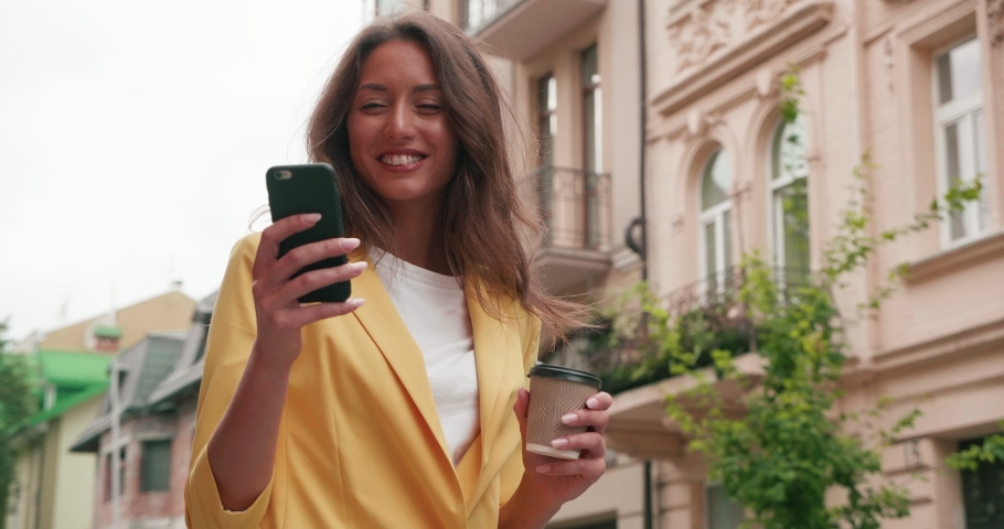 Pretty Young Woman Having Fun in the Town while walking with Mobile Phone in her Hands. Old Buildings in the Background. Smiling and satisfied girl holding cup with Hot Coffee to go