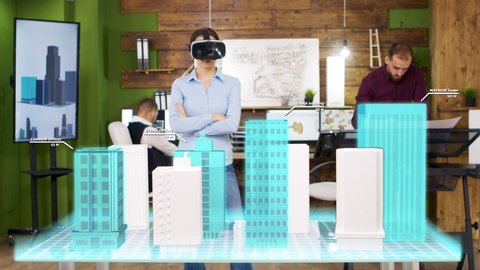 Architect woman wearing VR headset is looking at holographic projections of building a city. She rearranges the floors of a business tower स्टॉक वीडियो