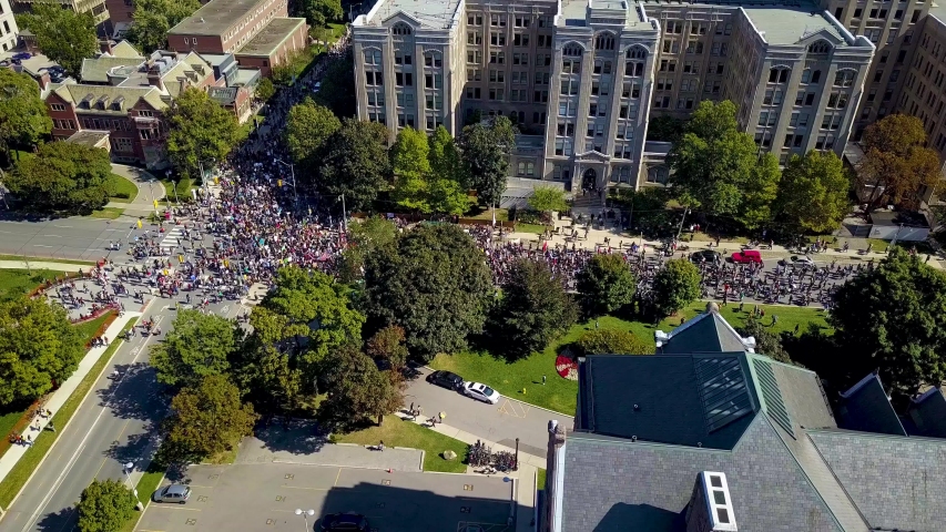 Huge crowd marches past old buildings in Toronto, aerial drone pan. Many people protest climate change by walking together on downtown city street, wide daytime exterior in 4k Royalty-Free Stock Footage #1045698460