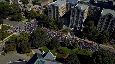 Large crowd marches by stone buildings in Toronto, aerial drone pan. Lots of people protest with democratic free speech by walking on downtown city street together, wide daytime exterior in 4k