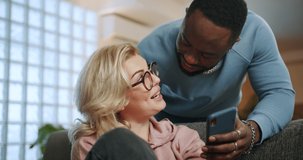 Affectionate Afro-American boyfriend sharing social media content with nice girlfriend, lovely couple leisure man communicating with woman from back holding mobile phone smiling at home living room