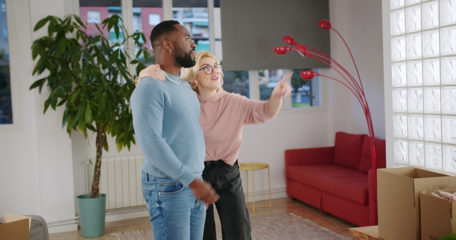 Glad mixed race couple discussing new flat interior of modern cozy living room together, happy blonde girlfriend hugging and kissing her lovey boyfriend just moved at home with positive feelings