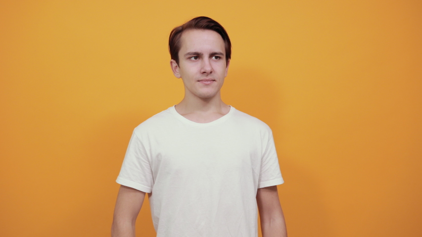 A young handsome guy grimaces and shows mimicry that he is eavesdropping. In white t-shirt on yellow background