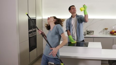 Two young people dancing together and emotionally singing using broom and cleaning tools as as microphones while cleaning in kitchen. Happy couple enjoying time, feel like rock stars
