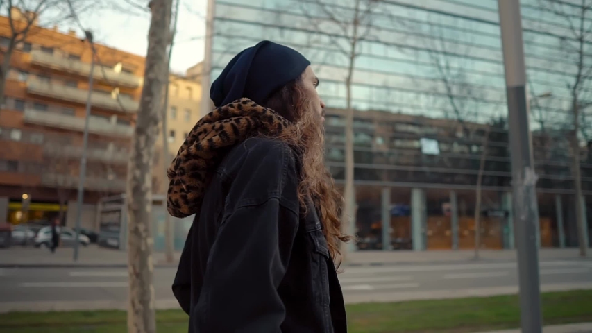 Portrait of long haired hipster walking on street in modern city in slow motion. He wears black jacket with leopard hoodie. Stylish man in sunset light