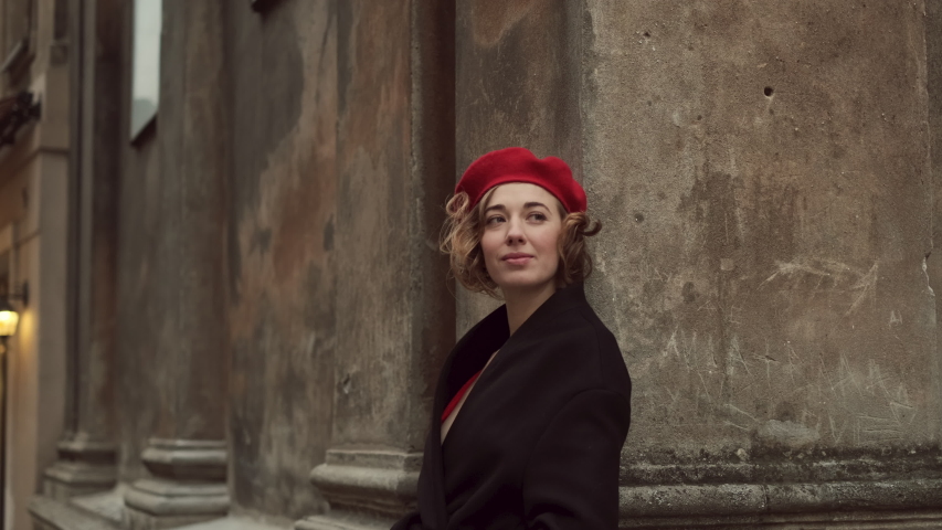 Romantic Young Woman Standing Touching Her Back Against An Ancient Wall In The City. Stylish Young Woman In Red Beret. Woman Smiling. Wind Moves Her Hair.