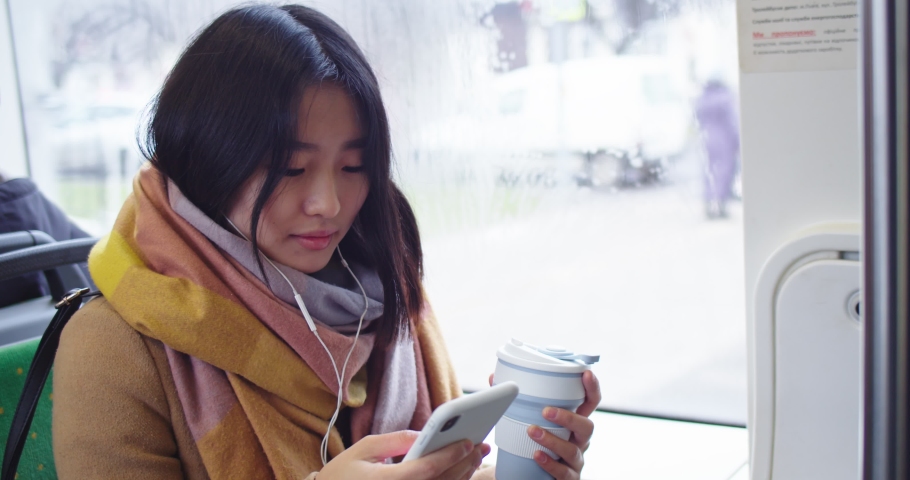 Asian young girl in headohones listening to the music and tapping on the smartphone while sipping hot coffee or tea and sitting in the tram or bus.