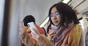 Asian young stylish and pretty girl in glasses and headphones tapping on the smartphone and listening to the music while standing in the tram or bus.