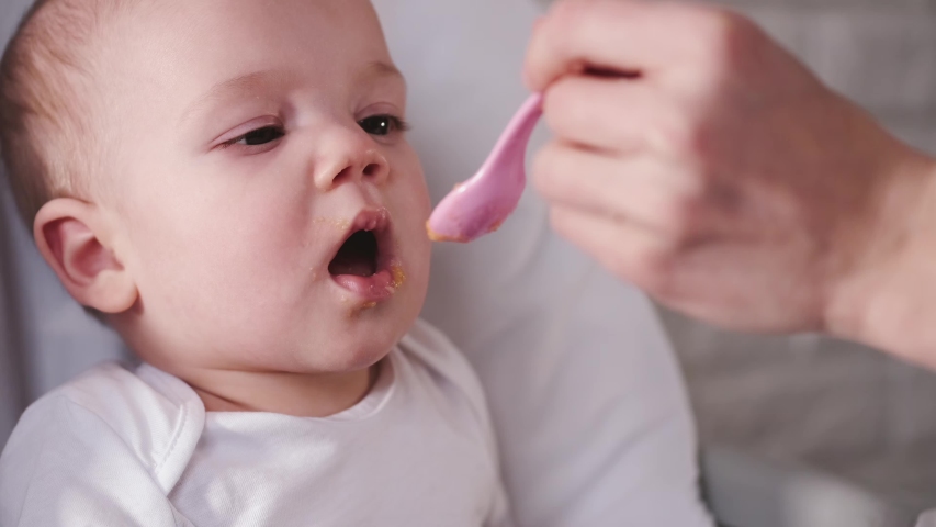 A baby is being fed by it’s mother with a spoon. The newborn is having a meal with food remains on it’s mouth.