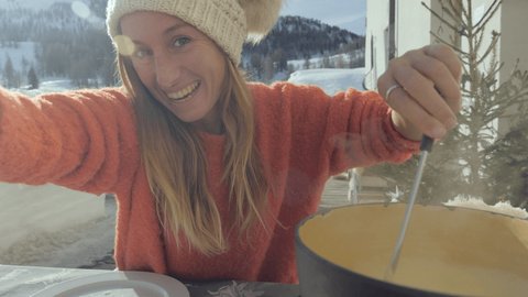 Fun video of young woman taking selfie while eating cheese fondue in the Swiss Alps in winter, mountains covered by snow, winter holidays concept. Food sharing communication 