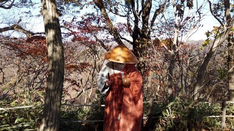 A Korean Buddhist Monk is on a mountain trail road surrounded by trees in Autumn, He is playing an instrument called a Moktak with a mallet as he bows his head