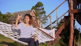 Asian woman making video call using smartphone with her boyfreiend  and  sitting in hammock at mornig time.