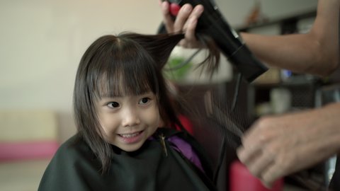 Hairdresser dries little girl hair with the hair dryer in the beauty salon. Asian little girl at hair salon. Little girl have happiness and enjoy in hair care. Handheld shot, Real life