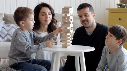 Family of four people is playing in board game with wooden tower together at home. Junior son is pulls out the brick and tower is crumbling