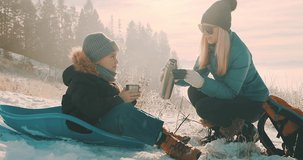 Happy mother and son drinking hot tea after fun in winter forest, beautiful snow landscape on sunny day. Beautiful mountain landscape view. 4K slow motion video.
