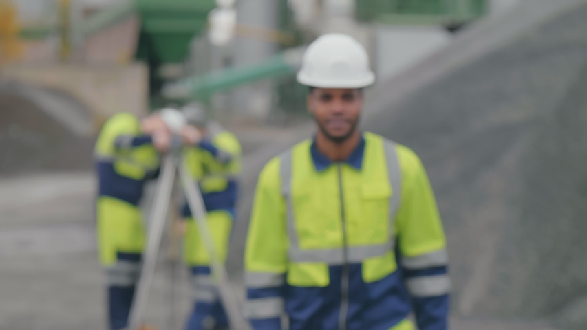 Close shot of positive worker walking towards camera. African geodesist in safety gear looking at camera and smiling, his colleagues using theodolite equipment on background at construction site. Royalty-Free Stock Footage #1045825918