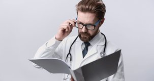 Concentrated medical expert in eyeglasses and white lab coat reading patient case while posing in studio with white background. Professional doctor examining medical history before treatment