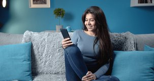 Happy young girl with long dark hair in casual clothing relaxing on grey couch and using mobile phone. Beautiful lady enjoying leisure time at home with modern design.