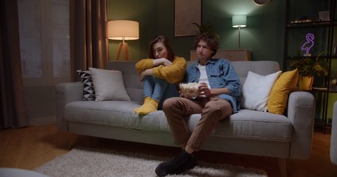 Young good looking Caucasian guy and girl watching interesting film on TV together with popcorn in the dark living room on the couch. Quarantine, pandemic, virus. Stay home