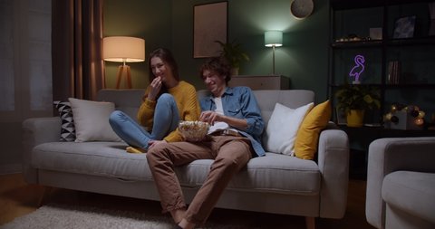 Young cheerful Caucasian couple sitting on the couch in the dark living room at night, eating popcorn and watching comedy movie on TV while laughing.
