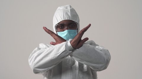 Portrait of an african-american man in protective suit, mask and glasses looking at camera. Showing crossed hands gesture. Stop signing. Studio, isolated on white background.