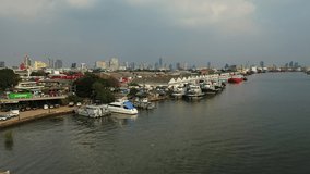 Aerial drone view of the Bangkok Port terminal facilities, cargo containers, boats and cargo ships. View from the river bank side, boat pier and the city skyline with skyscrapers in the background.