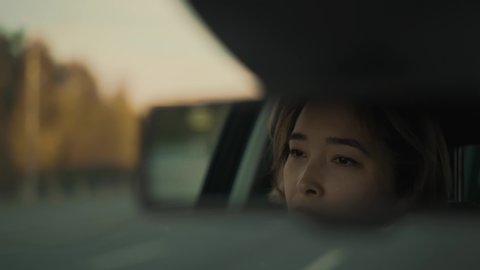 Handheld shot of serious asian woman in front view mirror while driving car on a high way. Woman in a car driving fast on the road during sunny evening