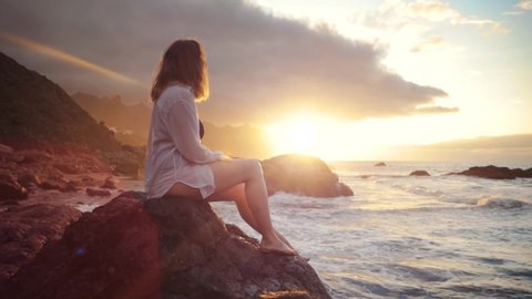 Side view of caucasian woman sitting on rock in sea enjoying relaxing sunset at the backview sunny evening nature travel ocean big waves outdoor breeze slow motion