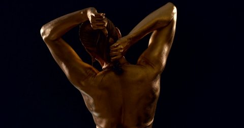 a sporty woman with raised muscles and Golden skin poses on a black background with her back to the camera. she holds her hair in her hands