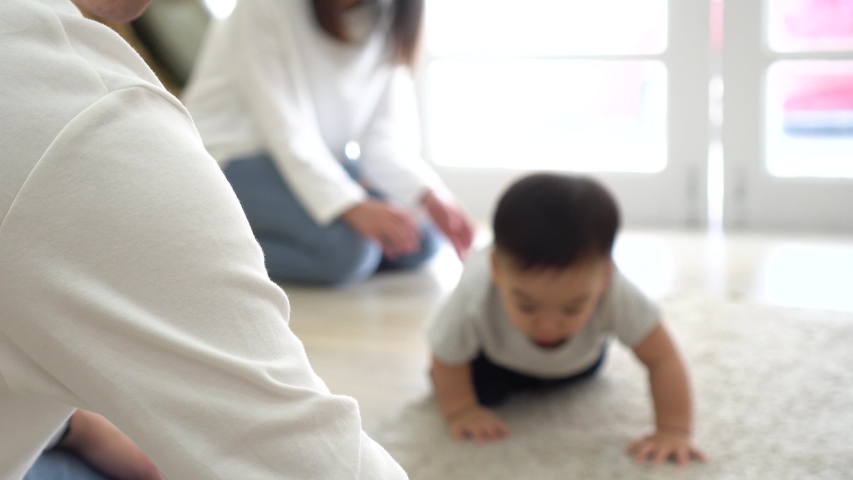 From above of Adorable Asian Infant boy looking at toy in hands of mom and crawling on carpet towards parents while family spending time together at home