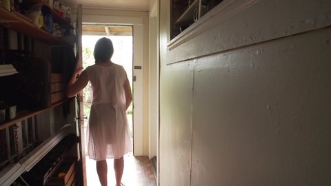 Wide shot of older an older woman in a night gown and slippers approaching a door in a dark hallway, then opening the door allowing bright daylight to spill in. shot at 240fps